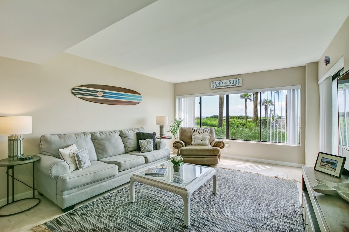 A light-filled living area features a comfortable sofa complemented by decorative pillows. A coffee table sits in front, with a subtle area rug beneath. Large windows provide views of the surrounding greenery, enhancing the sense of openness and connection to the outdoors.