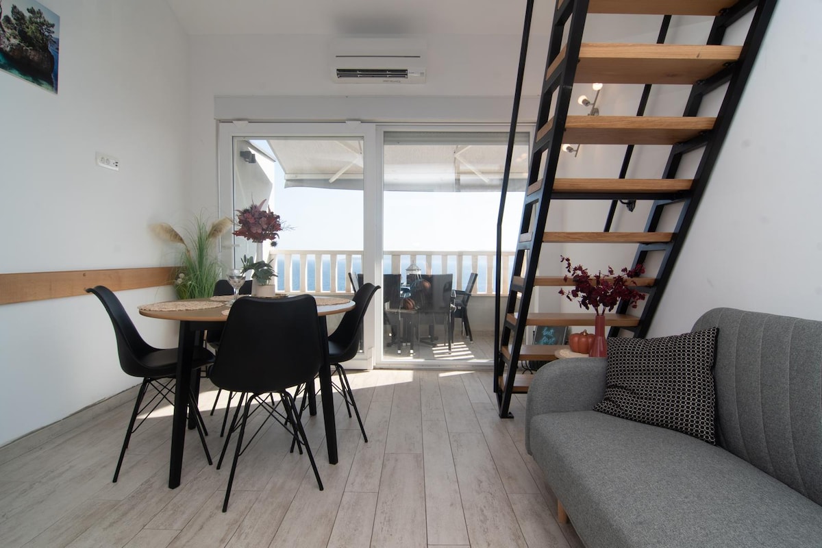 A dining area features a round wooden table surrounded by black chairs, with a scenic view accessed through large glass doors. A gray sofa is positioned nearby, and a staircase with wooden steps leads to an upper level. Natural light brightens the space.