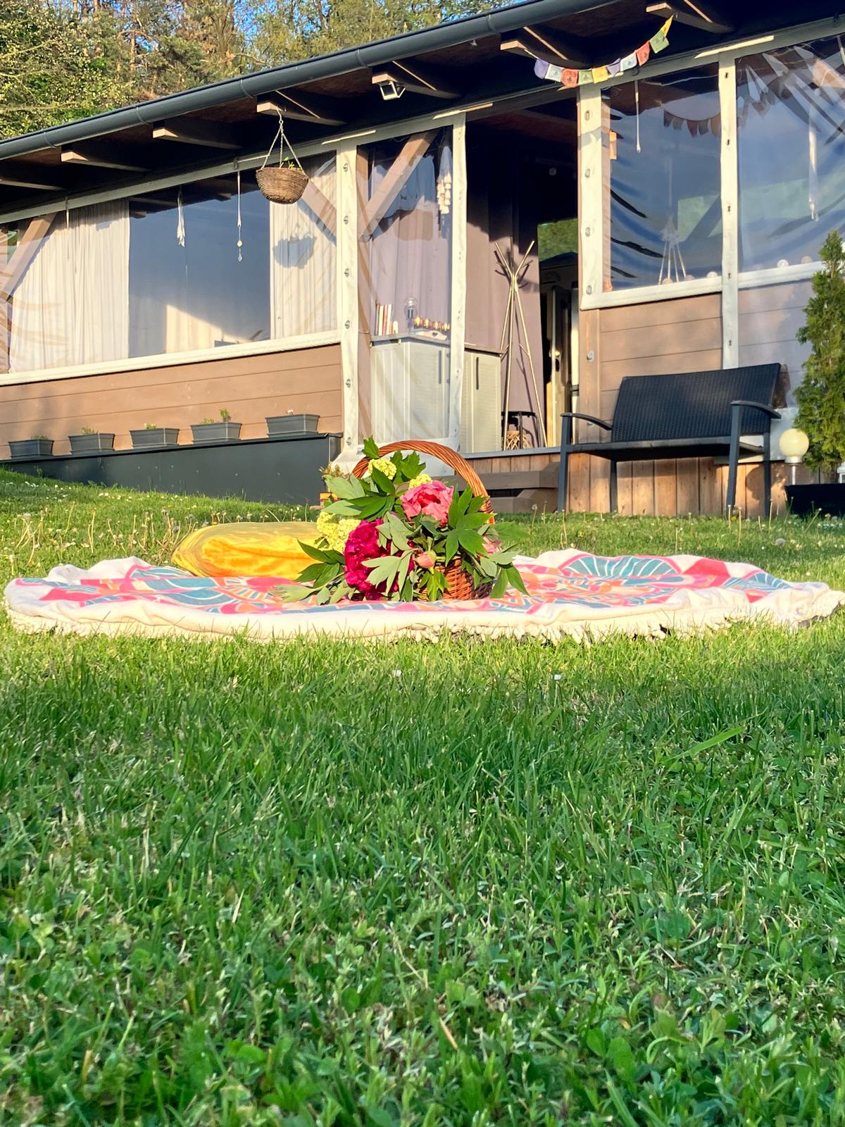 A picnic setup on a grassy area features a colorful blanket adorned with floral patterns, accompanied by a woven basket and decorative foliage. The background includes a wooden structure with large windows and a seated area, surrounded by potted plants and natural greenery.