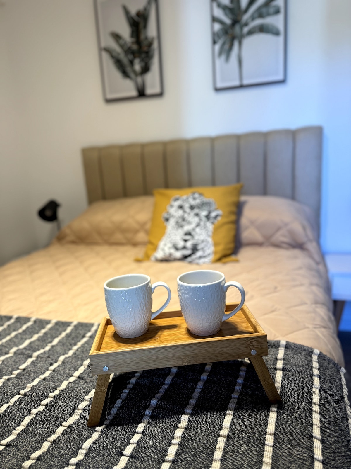 A cozy bedroom is highlighted by a spacious bed with a neutral-toned quilt. Two cups sit on a wooden tray, resting on a folded blanket. Above the bed, wall art featuring palm leaves complements the relaxed ambiance.