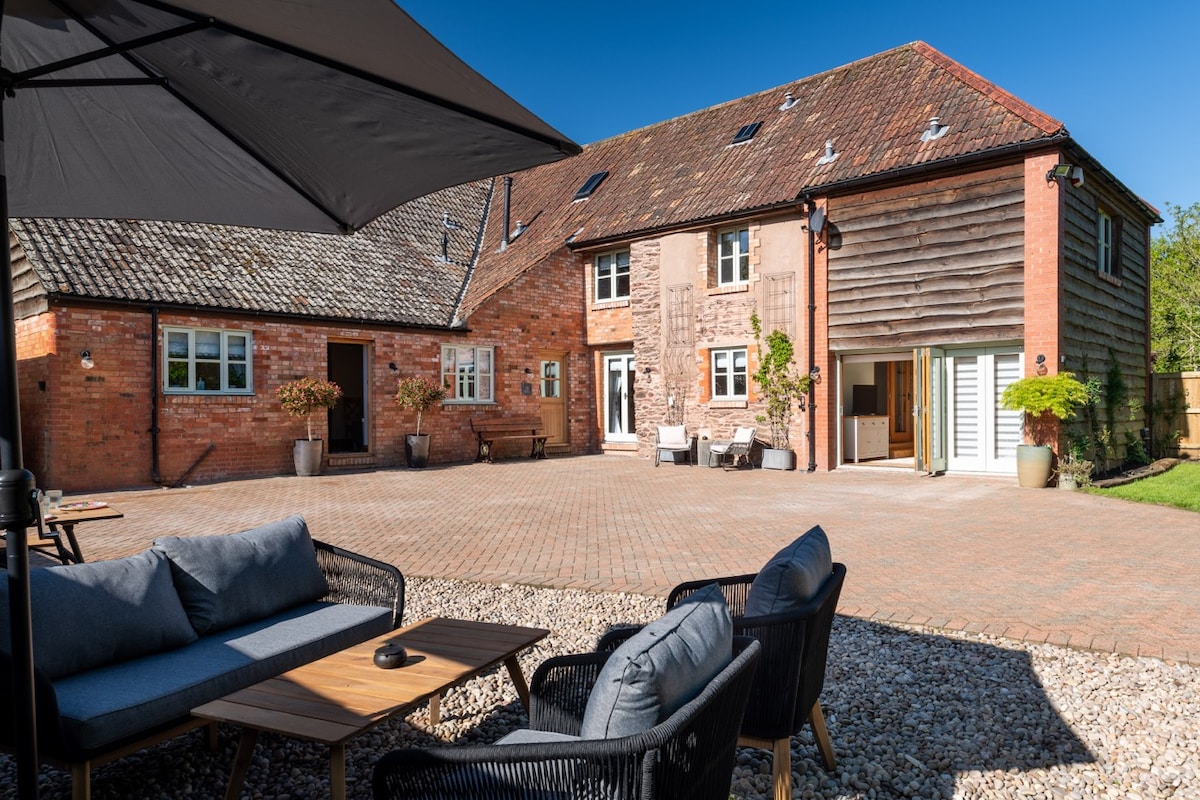 An outdoor seating area is presented with comfortable chairs and a wooden table. The spacious cobblestone courtyard is framed by the barn's rustic brick and wooden exterior, complemented by potted plants. Sunlight brightens the scene, creating a welcoming environment for relaxation.