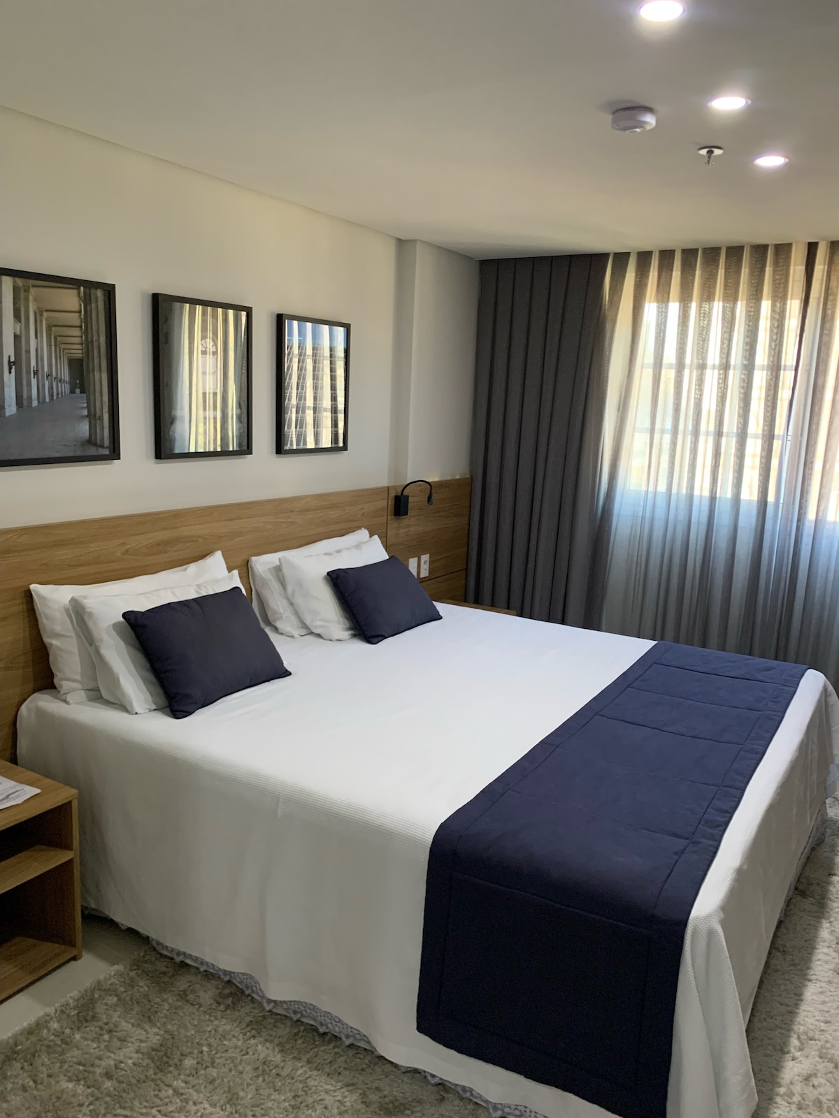 A well-appointed bedroom features a spacious bed with white linens and navy accents. Natural light filters through sheer curtains, highlighting three framed mirrors on the wall. A bedside table stands to the left, maintaining an organized and modern aesthetic.