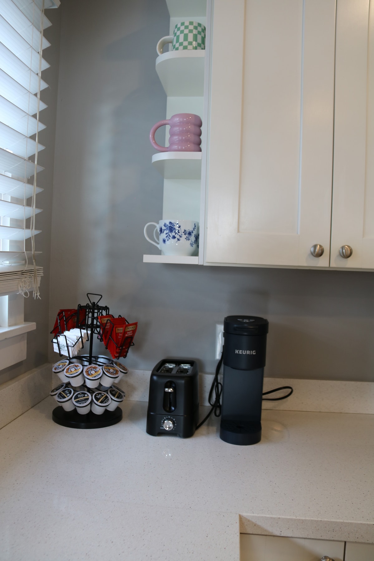 A modern kitchen corner is shown, featuring a Keurig coffee maker and a toaster on a light-colored countertop. Above, open shelves display colorful mugs and dishes, adding a touch of charm. A coffee pod holder sits nearby, neatly organized.