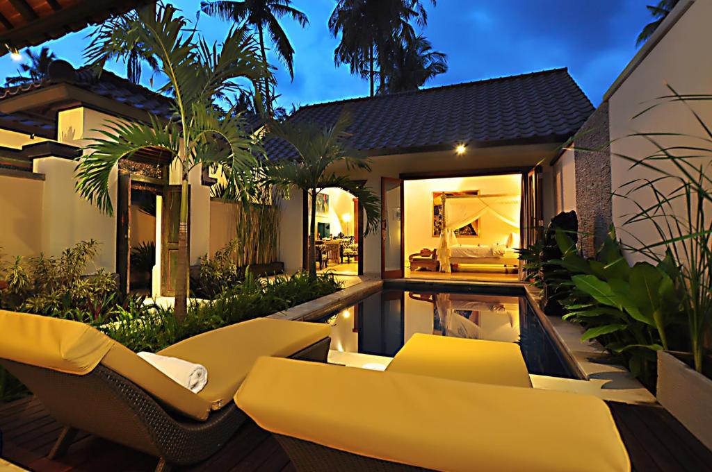 A private outdoor area is highlighted by two sun loungers, accented with yellow cushions, positioned beside a serene pool. Lush tropical plants surround the space, and the villa's living area can be seen through large glass doors, creating a cohesive indoor-outdoor environment.
