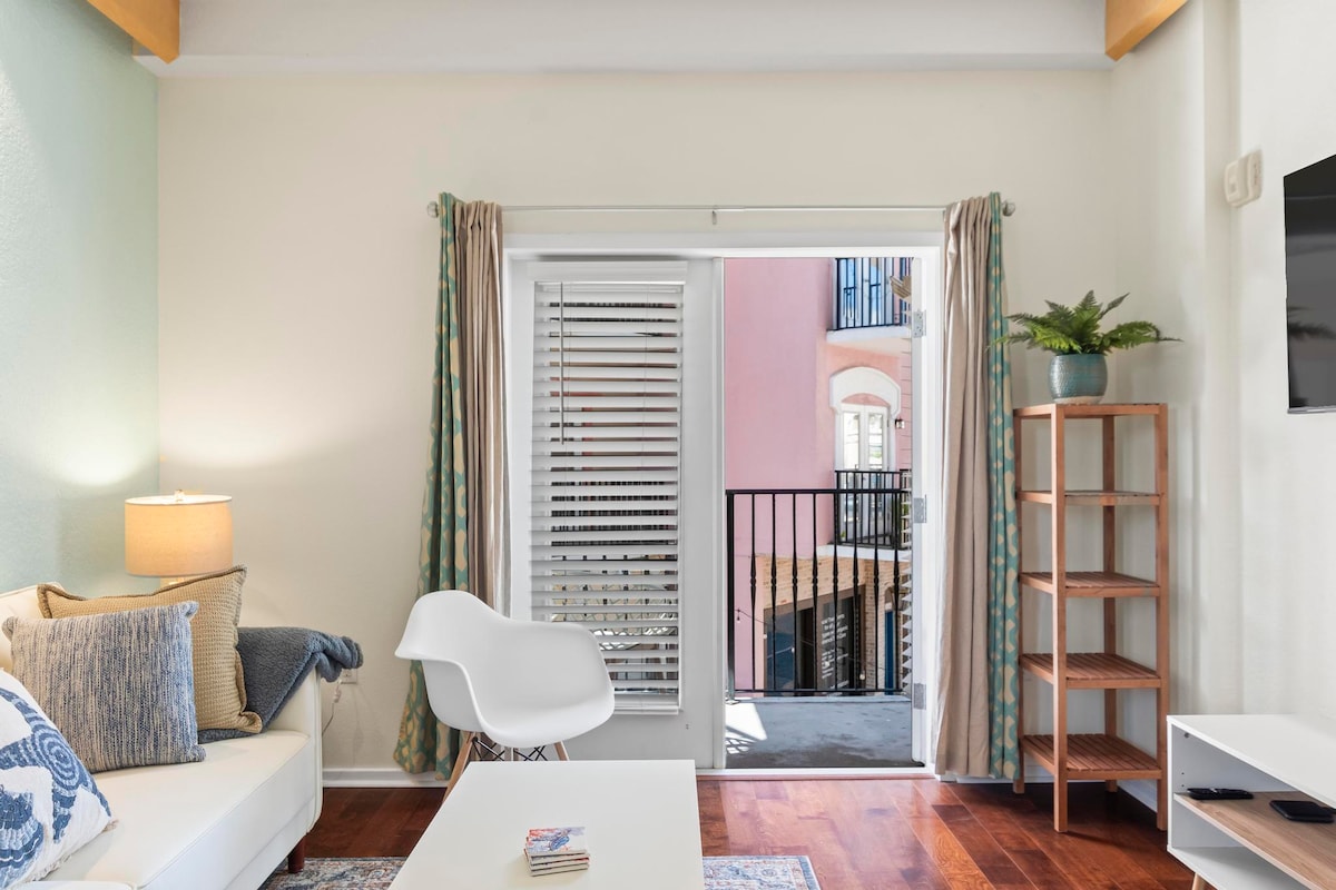 The living area features a light-colored sofa and a small coffee table. Sliding glass doors allow natural light to enter, offering a view of the balcony and courtyard below. Decorative curtains frame the entrance, and a potted plant adds a touch of greenery.