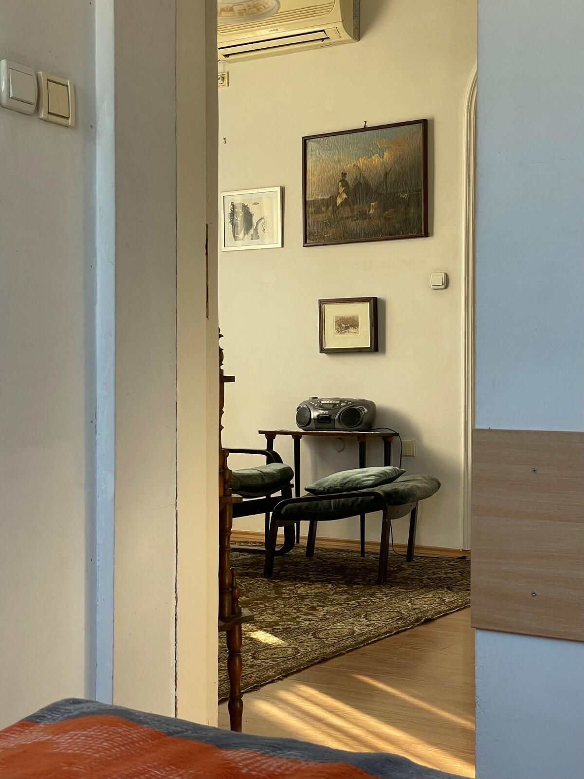 A glimpse into a cozy seating area is provided, showing two green upholstered chairs beside a small table. Wall art adorns the light-colored walls, and a decorative rug lays on the floor, adding warmth to the space. Soft lighting enhances the inviting atmosphere.