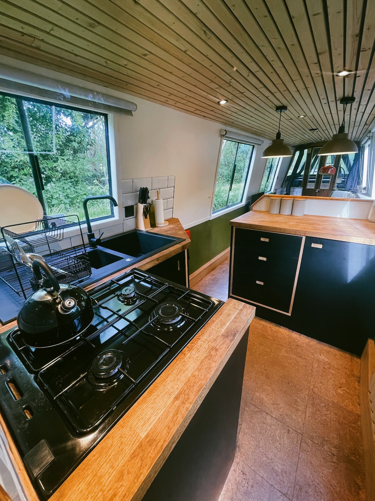 The galley features a four-ring gas hob with a kettle on top, complemented by a wooden countertop and a black sink. Natural light flows through nearby windows, and a green wall adds a touch of color. Functional shelving and storage units are visible, emphasizing a well-organized layout.