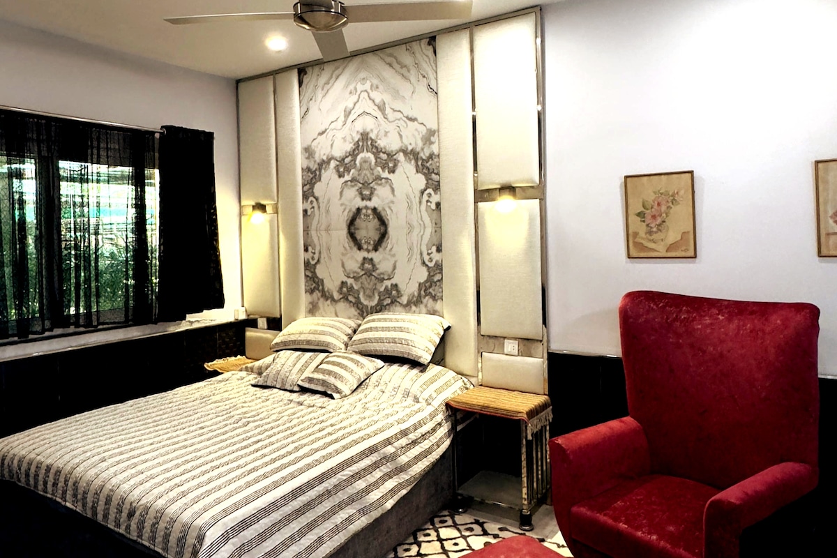 A spacious bedroom features a large bed with striped bedding, complemented by a patterned headboard. A vibrant red chair adds a splash of color. Natural light filters through the window, which is framed by dark curtains, creating a balanced contrast in the room's design.
