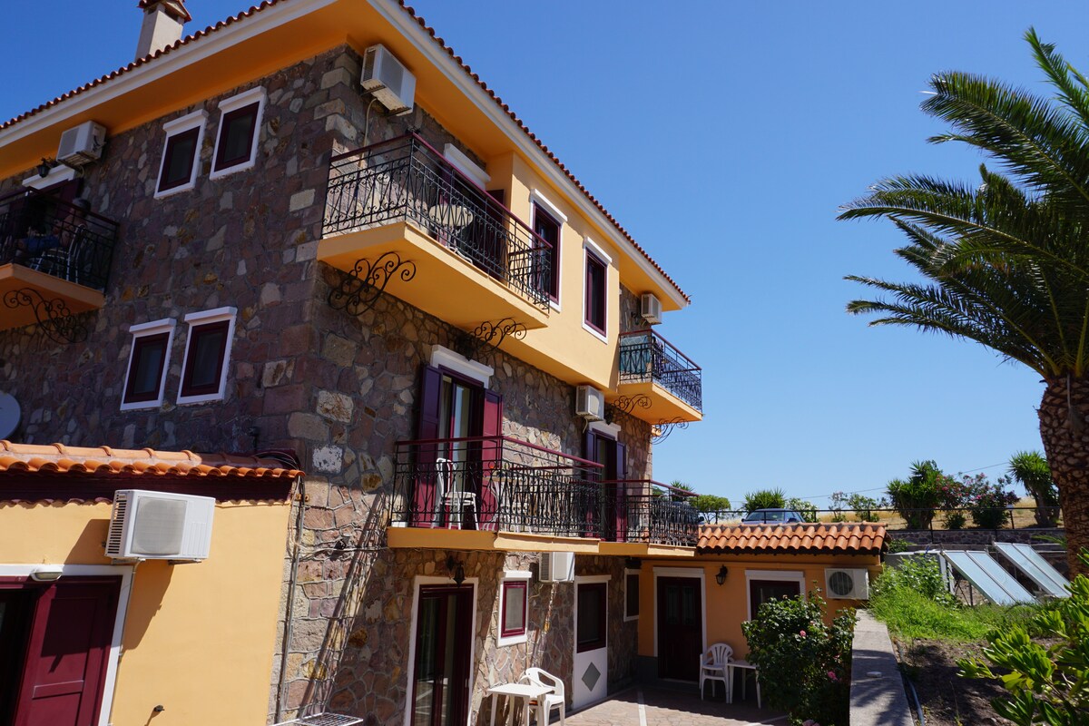 A multi-level building is constructed with stone walls and features balconies with wrought iron railings. Air conditioning units are visible on the exterior. Surrounding landscaping includes palm trees and green shrubs, contributing to a relaxed outdoor atmosphere.