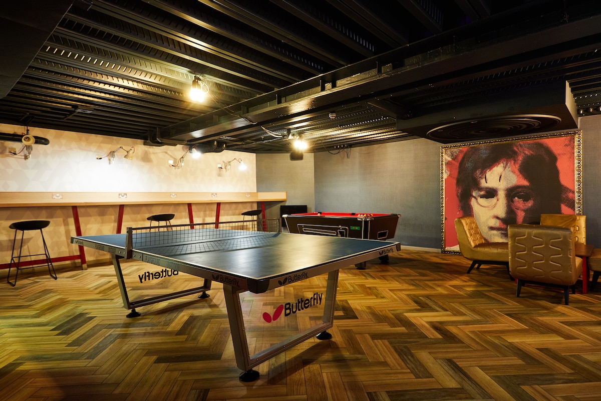 A game area is featured with a ping pong table at the center. To one side, bar stools line a counter, while a foosball table and comfortable seating are visible in the background. Warm lighting enhances the modern design of the space.