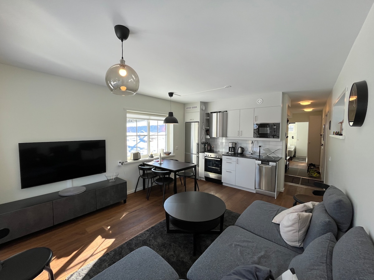 A modern living space features a comfortable sectional sofa positioned next to a wall-mounted television. A dining table for four sits nearby, with a kitchenette equipped with stainless steel appliances and ample counter space. Natural light floods the area through large windows.