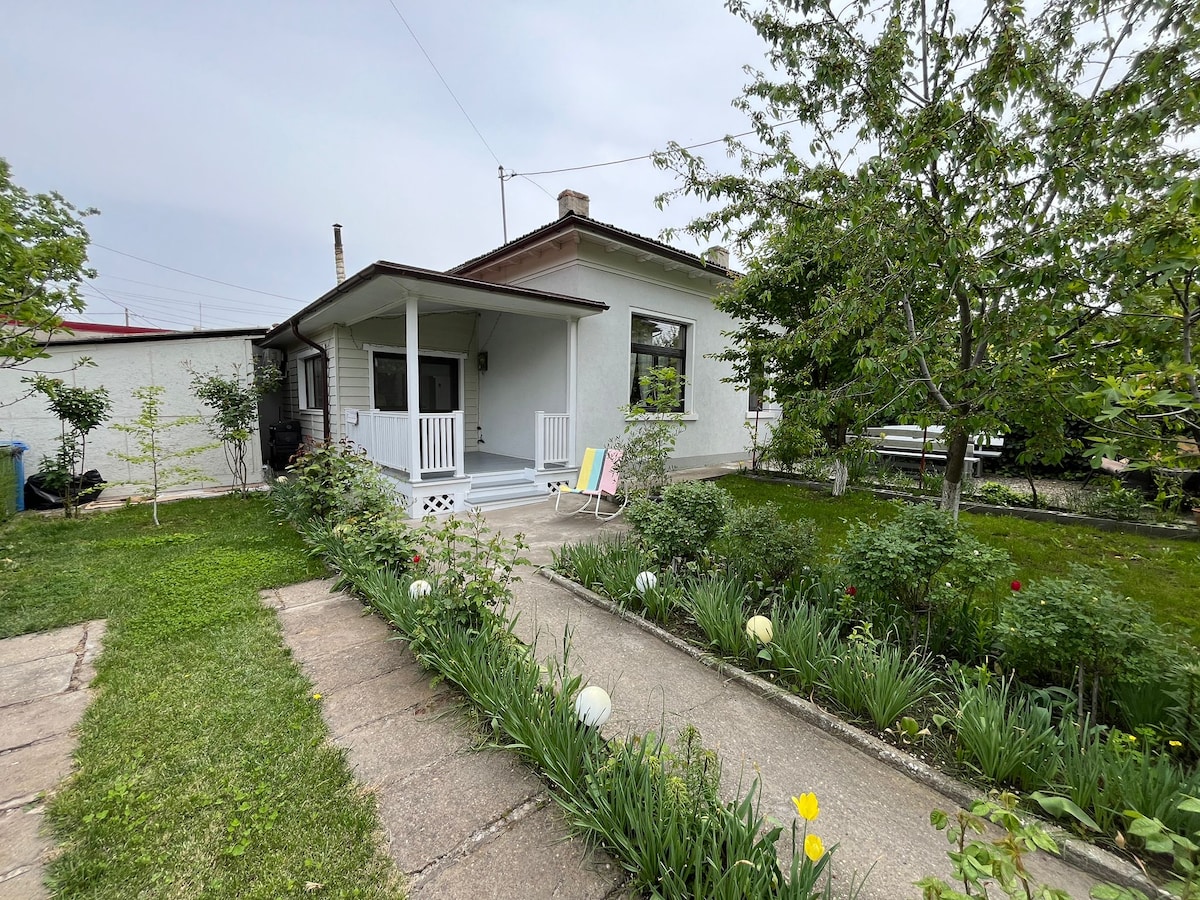 A charming single-story house is set among a well-maintained garden featuring a variety of plants. A porch with seating is visible, welcoming guests. A paved walkway leads through lush greenery, creating an inviting entrance to the home.