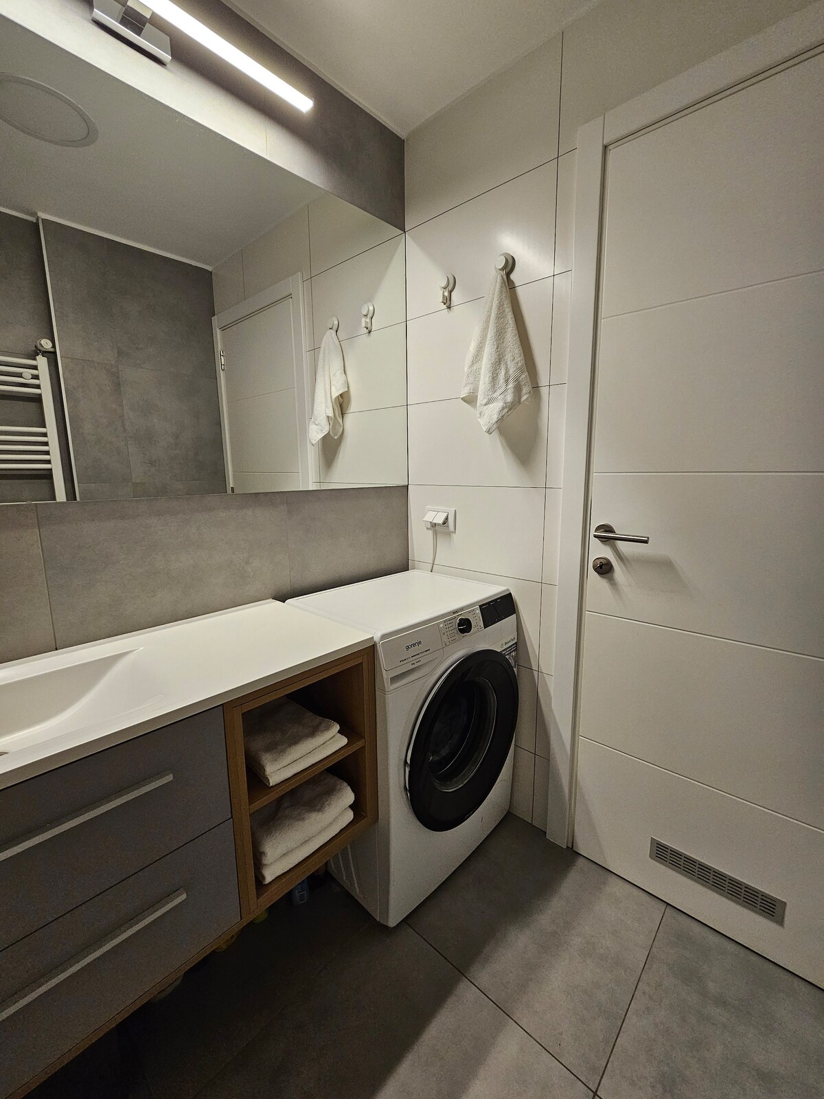 A modern bathroom is displayed, featuring a sleek vanity with ample counter space and a mirror above it. A washing machine is integrated into the cabinetry, and a linen cabinet holds neatly stacked towels. Soft lighting highlights the clean, tiled surfaces.