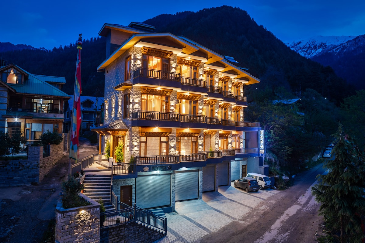 The exterior of the property is highlighted by warm lights illuminating the building at dusk. The two-story structure features multiple balconies with wooden railings, surrounded by lush green landscaping and distant snow-capped mountains offering a peaceful ambiance.