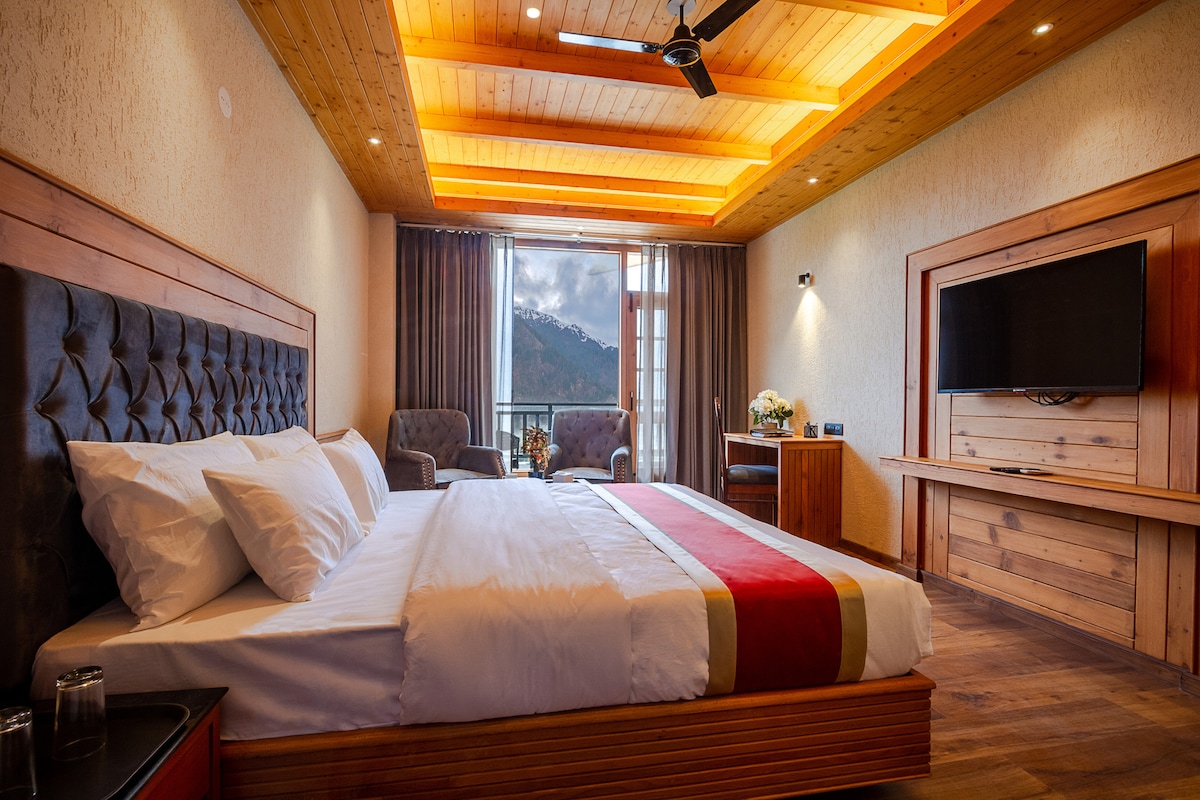 A spacious room is furnished with a large bed, accented by a red runner and crisp white linens. Two armchairs are positioned nearby, with a wooden desk hosting a decorative vase. Natural light streams through large windows revealing mountain views, while soft lighting complements the wooden ceiling.
