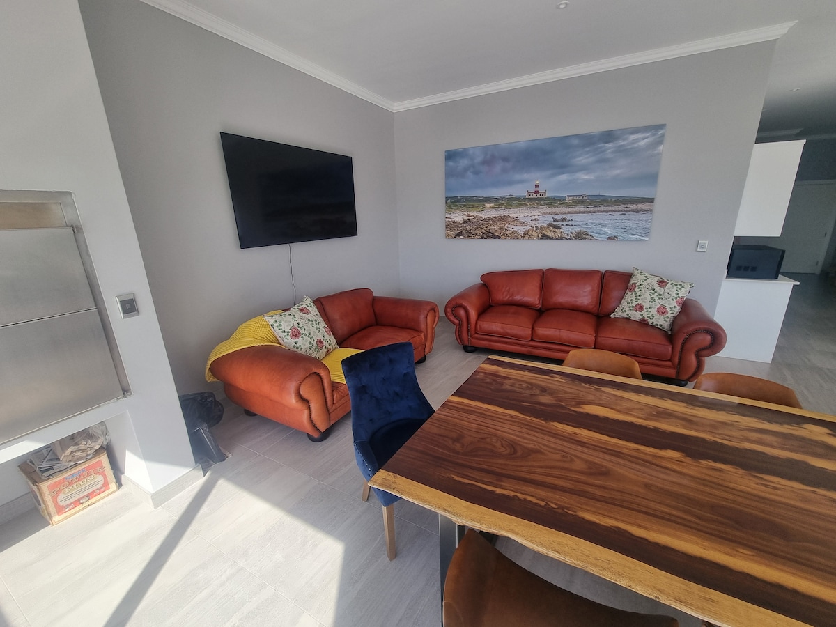 A warm living area features two brown leather sofas, one adorned with a colorful throw pillow. A wooden dining table occupies the space, surrounded by chairs in various colors. A large TV is mounted on the gray wall, complemented by a coastal artwork above the sofas.