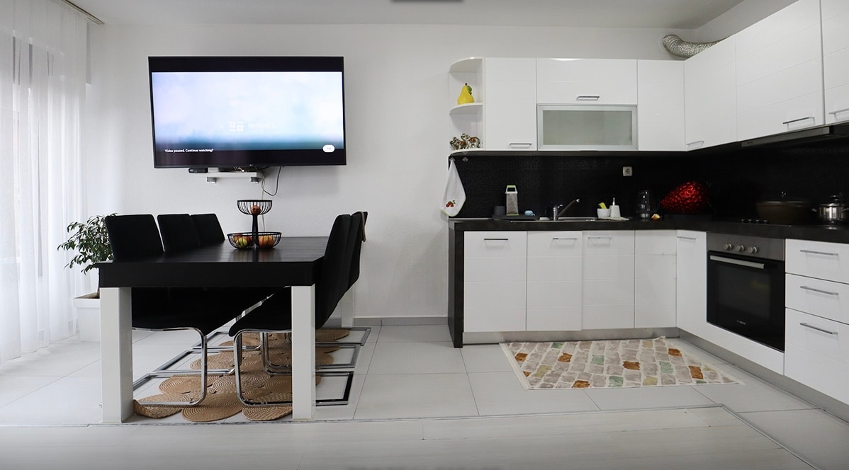 A modern kitchen and dining area feature sleek white cabinetry and a dark countertop. A black dining table is surrounded by six chairs, with a wall-mounted television above. A small plant adds a touch of greenery, and a textured rug lies on the floor near the kitchen area.