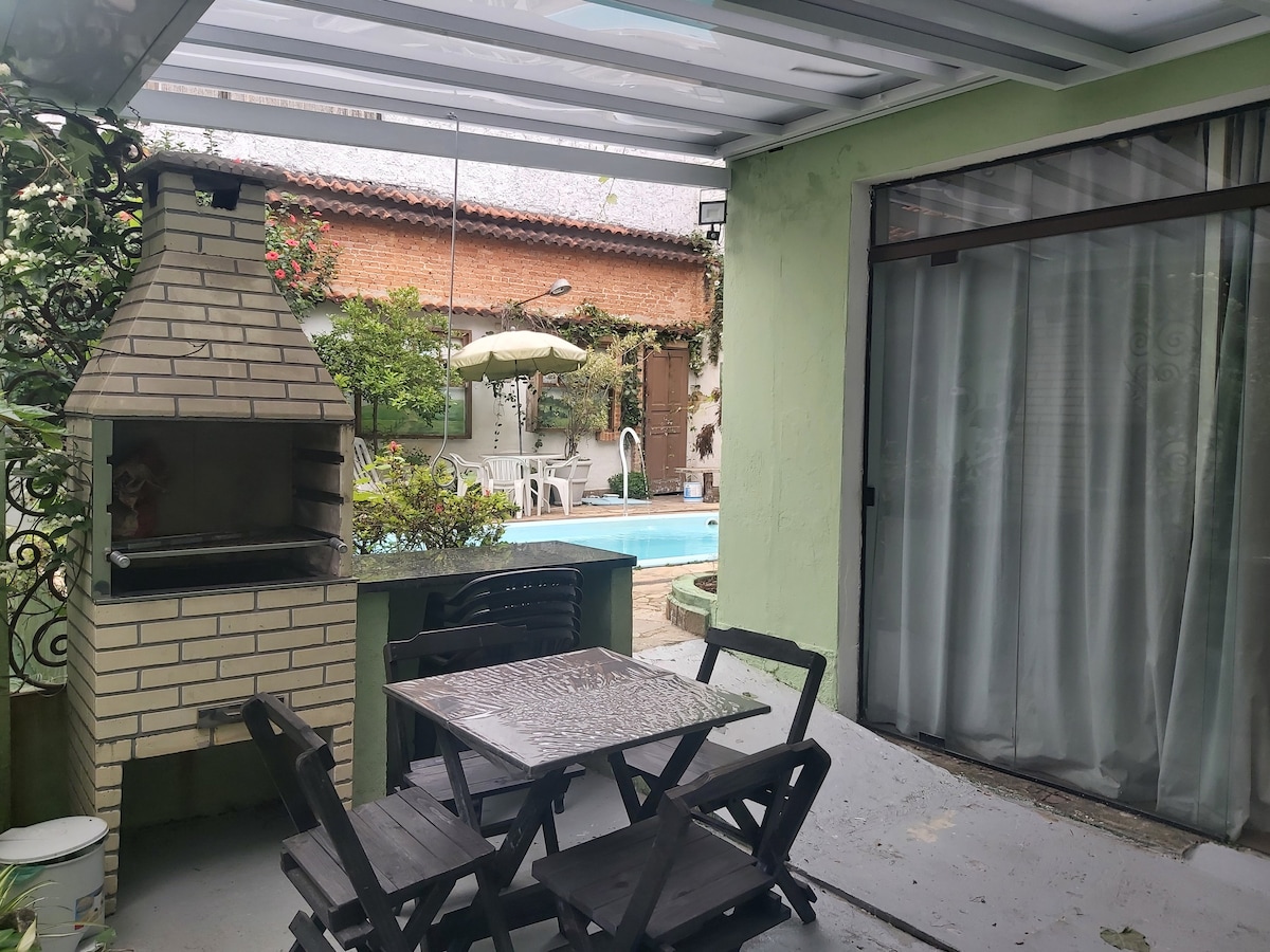 A shaded outdoor seating area features a wooden table with four chairs under a glass-covered patio. A brick barbecue grill stands nearby, with lush greenery and a swimming pool visible in the background. Light filters through sheer curtains from an adjacent room.