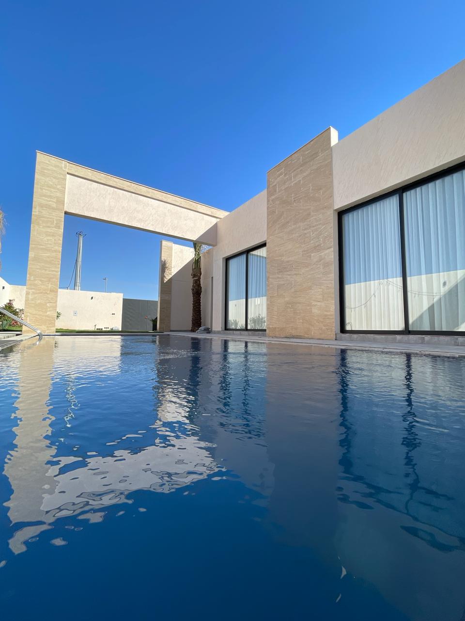 A contemporary villa is reflected in the still waters of an inviting pool. The structure features large glass windows, allowing natural light to fill the spaces, while minimalistic architectural elements enhance the serene outdoor setting against a clear blue sky.
