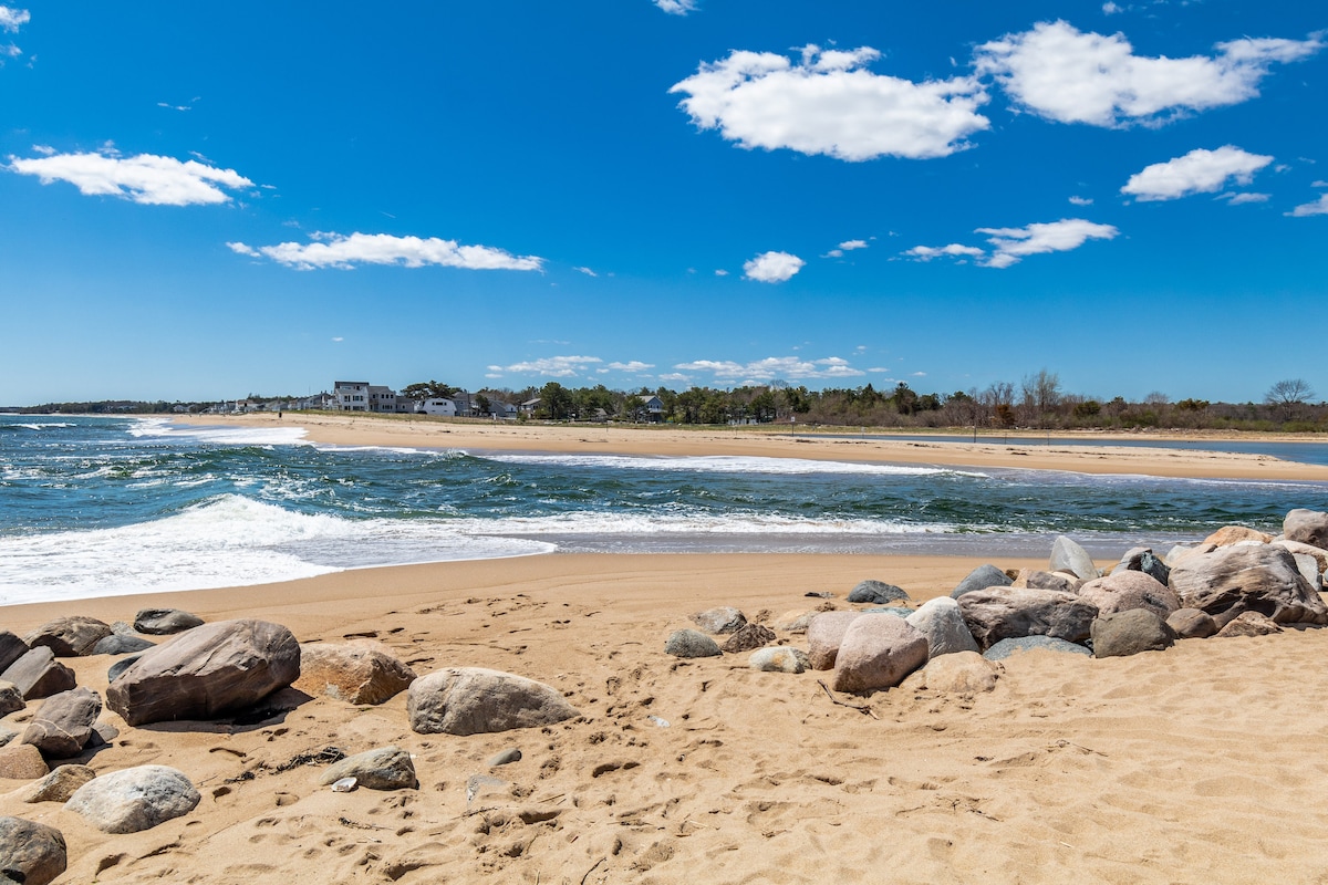 A sandy beach is framed by a gentle curve, where the Atlantic Ocean meets the shore. Smooth rocks are scattered along the sand, with clear blue skies and fluffy white clouds overhead. A glimpse of distant buildings and greenery can be seen along the horizon.