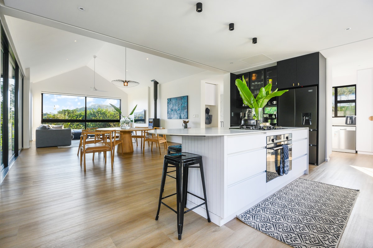 A spacious open-plan kitchen and living area is showcased, featuring a large island with seating, modern appliances, and a dining table set for gatherings. Expansive glass doors provide a view of the lush surroundings, allowing natural light to fill the space.