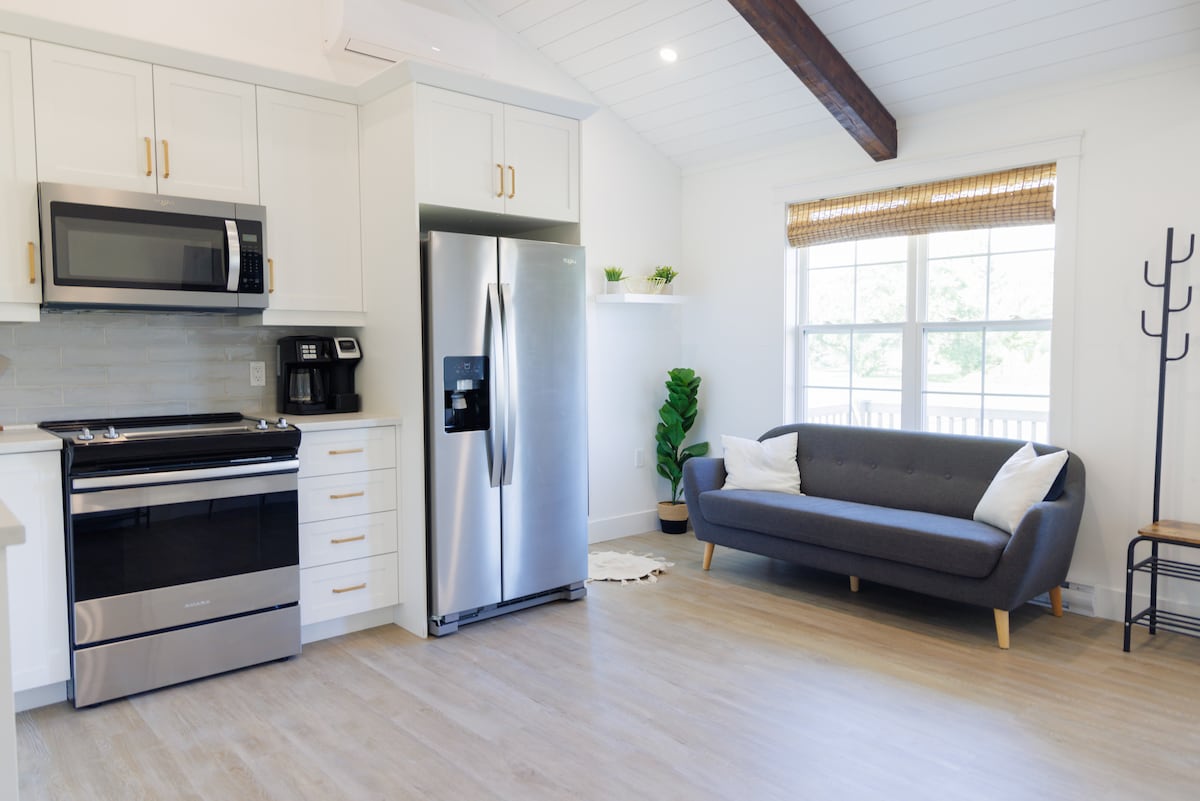 A modern kitchen features stainless steel appliances, including a microwave and a refrigerator. A sleek, gray sofa is positioned beside a window adorned with natural light and a woven shade. Indoor greenery is present, and the light wood floor adds to the bright atmosphere.