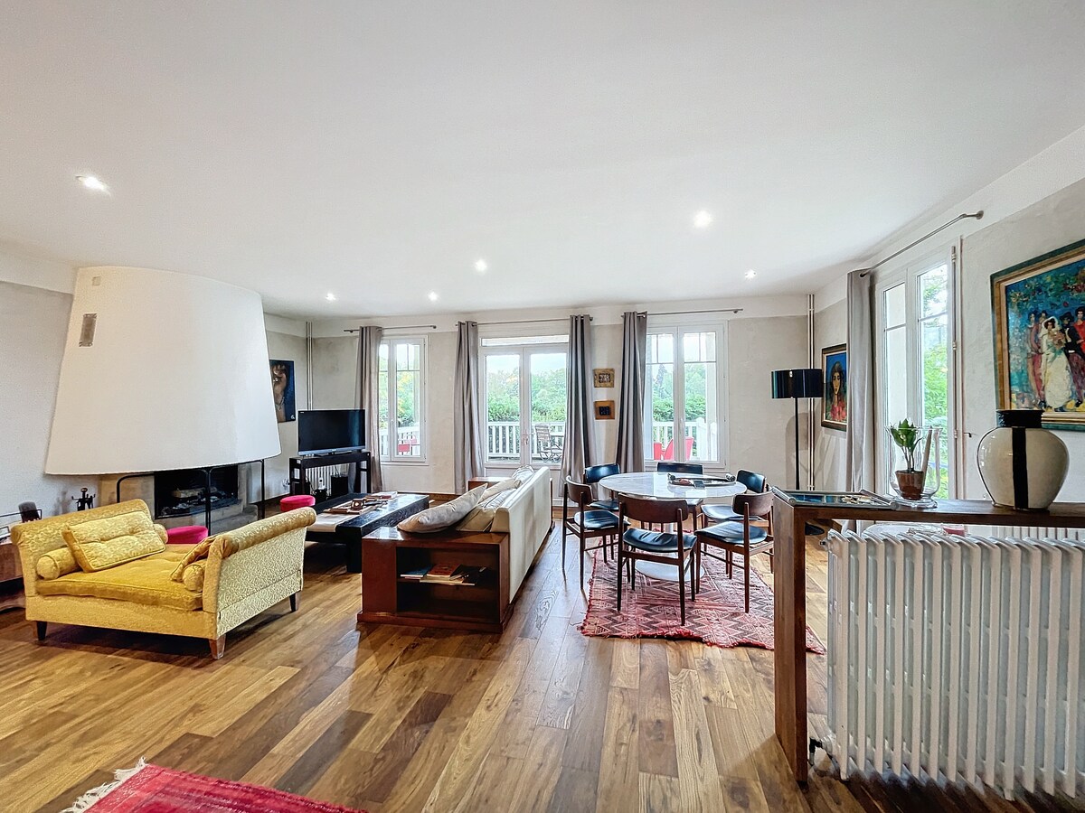A spacious living area is presented with large windows that allow natural light. A modern fireplace is situated near comfortable seating, and a dining table is set for six. Art pieces adorn the walls, enhancing the room's ambiance. Wooden flooring adds warmth to the space.