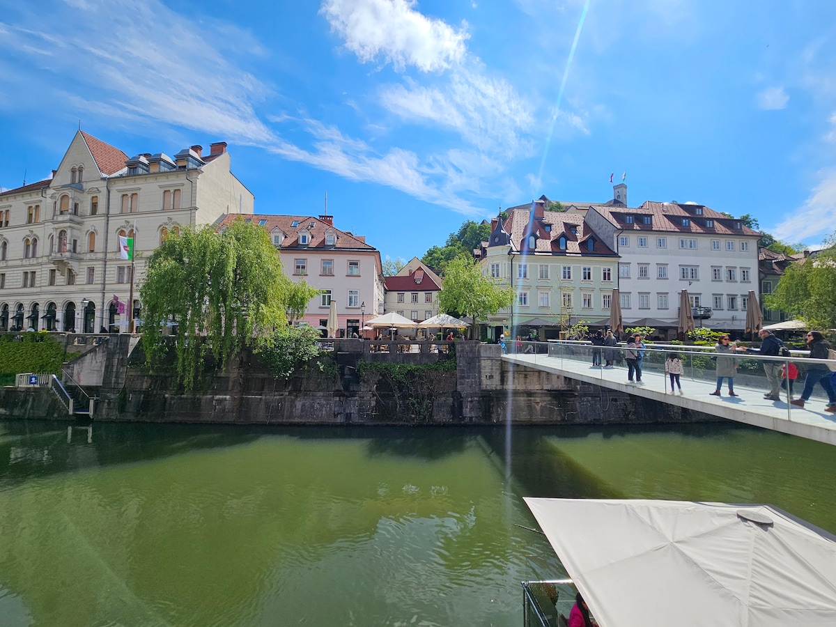 A wide view of a serene riverside scene along a flowing waterway, with various historic buildings lining the banks. People can be seen strolling along a pedestrian bridge connecting the two sides, underscored by a clear blue sky.