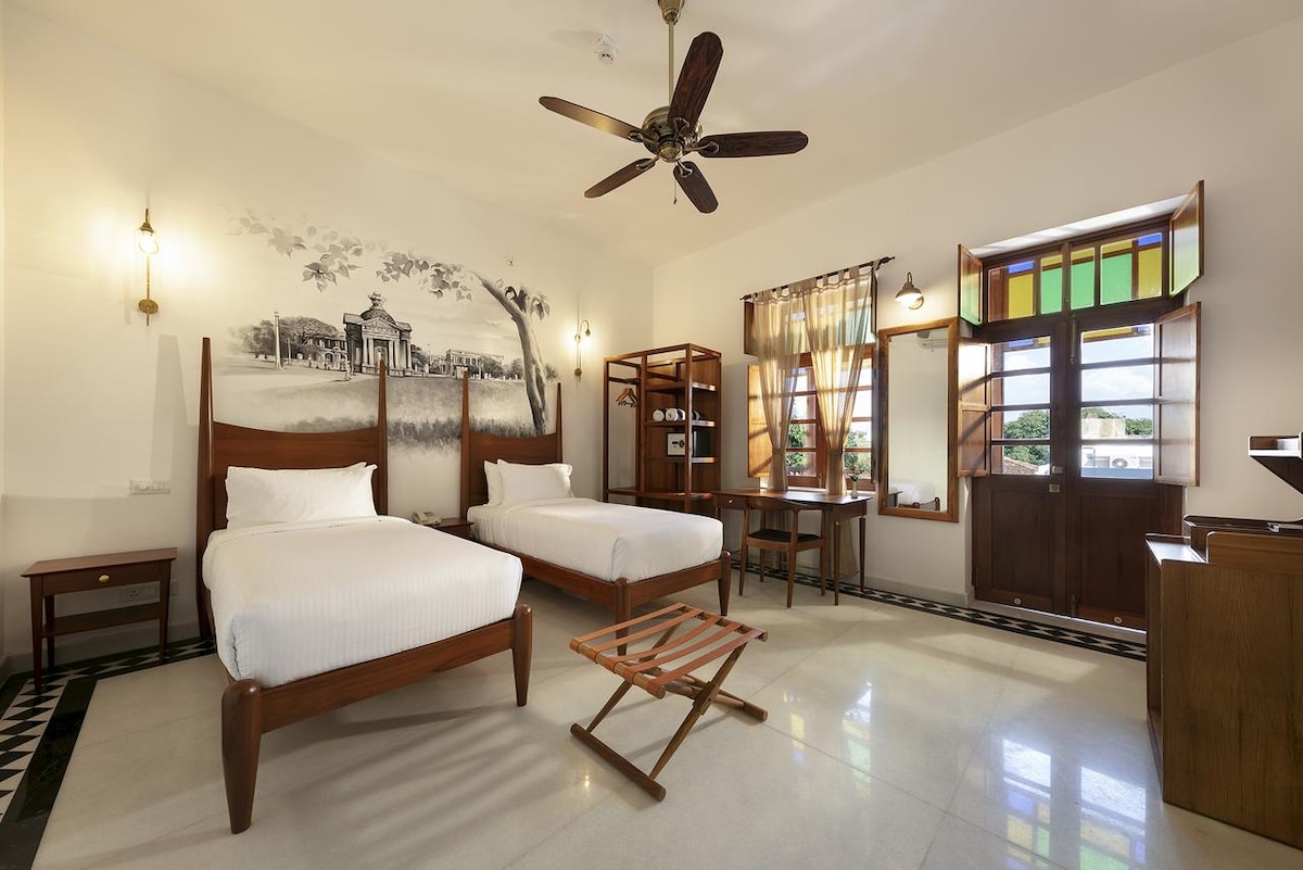 A spacious room features two beds placed against a mural depicting a scenic view. Natural light enters through large windows, adorned with wooden shutters. A wooden table and a folding chair are positioned beside the beds, while a ceiling fan hovers above the room.