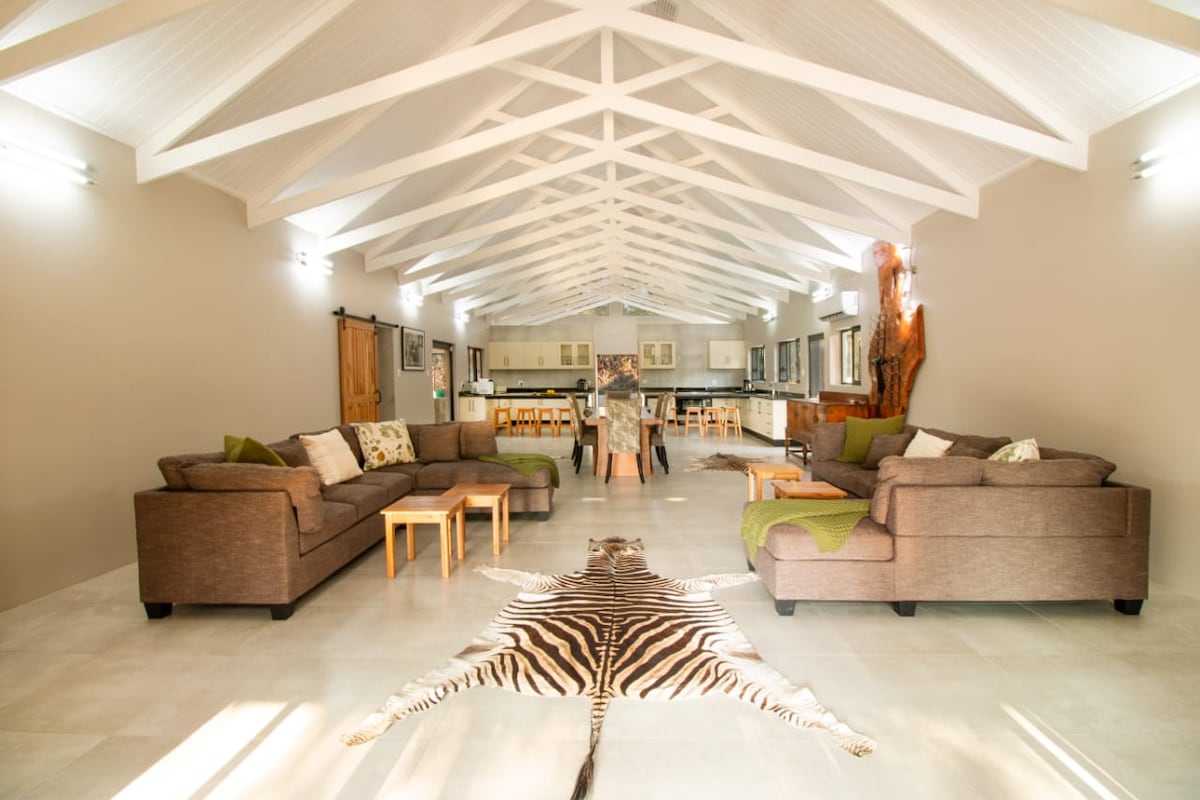 A spacious living area showcases high ceilings with exposed beams. Comfortable sofas are arranged around a central area, featuring earthy tones and decorative cushions. A zebra-patterned rug extends across the floor, leading towards a kitchen area with wooden stools and a dining table in the background.