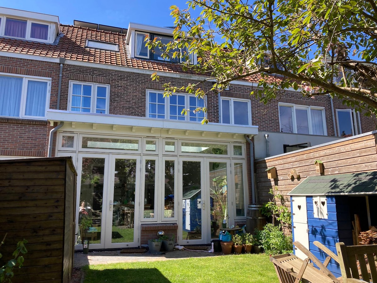 The image displays the rear view of a recently renovated family home, characterized by large glass windows that invite natural light. A well-kept garden, featuring various plants and a playhouse, is visible in the foreground, along with a wooden seating area.