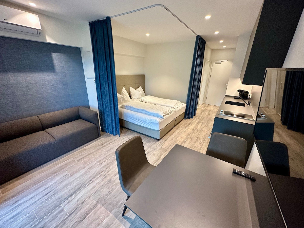 An open-plan apartment showcases a spacious sleeping area with a large bed. A dark grey sofa and a dining table with chairs occupy the living space. Light-colored wood flooring enhances the minimalist design. Floor-to-ceiling curtains frame the windows, contributing to a serene ambiance.