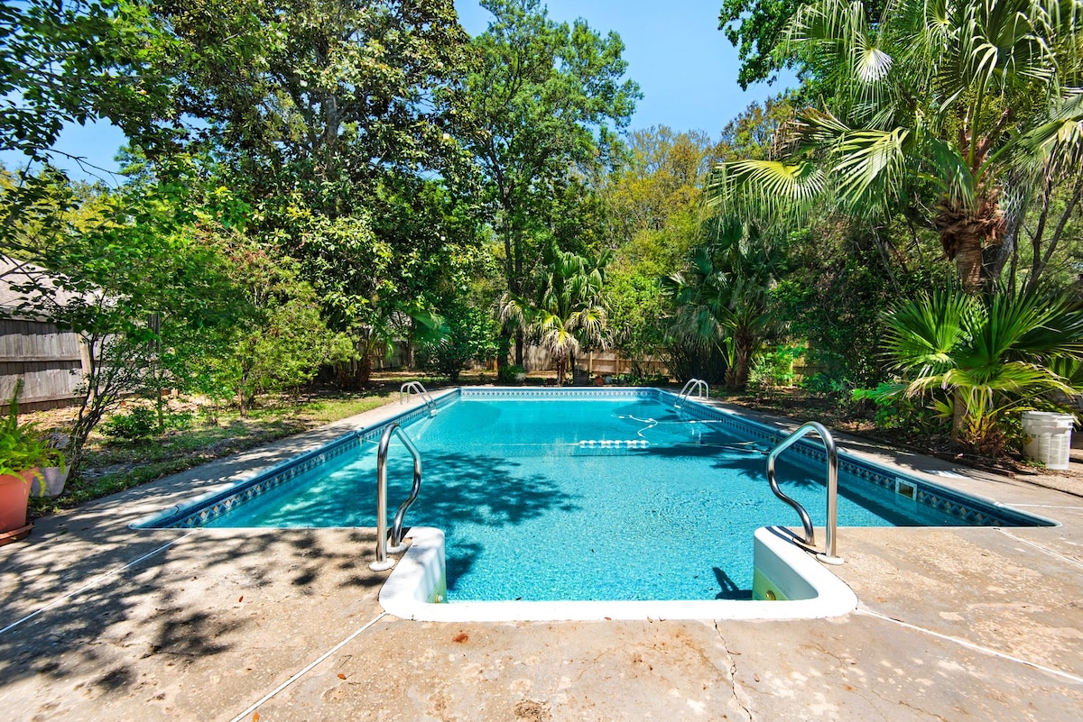 A spacious outdoor pool is framed by lush greenery and tall palm trees, creating a private oasis. Steps lead into the clear blue water, with a ladder on the opposite side for easy access. Sunlight reflects off the surface, enhancing the serene surroundings.