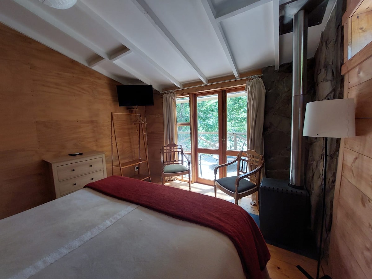 A cozy bedroom features a spacious bed draped with a soft blanket. Natural light enters through large windows, highlighting wooden walls and a stone chimney. Two wicker chairs and a small dresser complement the decor, creating a comfortable, inviting space.