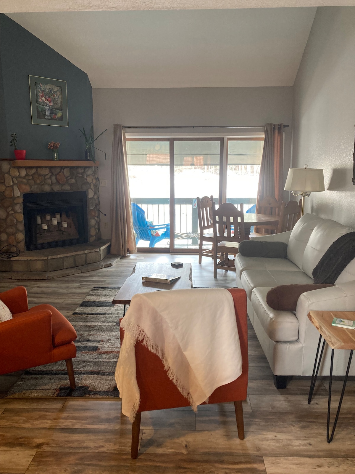 A cozy living area features a white sofa and two orange armchairs, positioned around a wooden coffee table. A stone fireplace serves as a focal point, while large glass doors lead to a balcony with outdoor seating. Natural light fills the space through the windows.