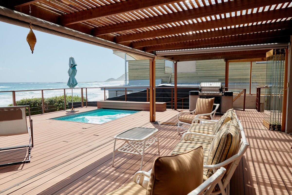 A spacious patio is equipped with comfortable seating and features a solar heated swimming pool. A barbecue grill is visible, alongside an outdoor dining area. A beach umbrella offers shade, while ocean waves can be seen in the background, creating a serene coastal setting.