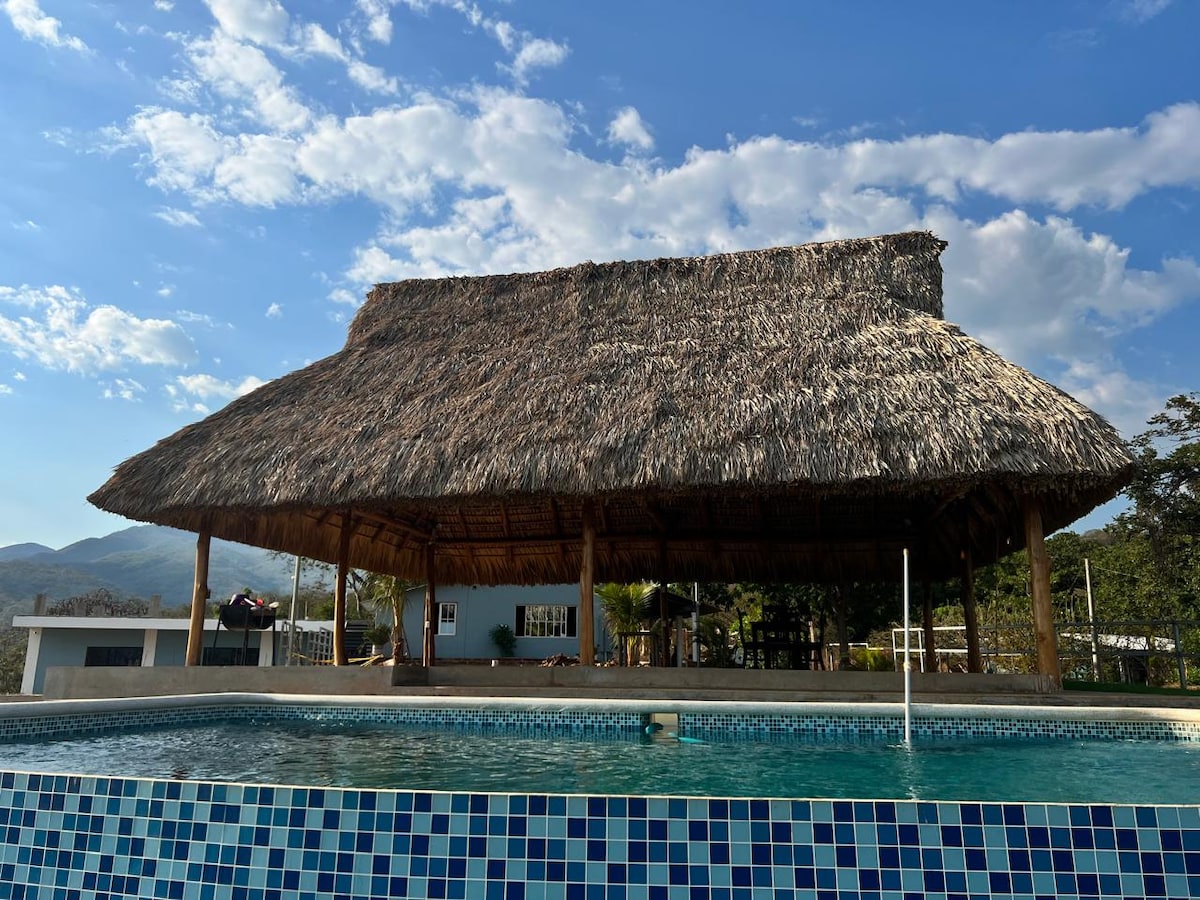 An open-air pavilion with a thatched roof provides shade near a rectangular pool. The pool is tiled with blue mosaics and reflects the sky. Lush greenery is visible in the background, along with distant mountains under a partly cloudy sky.