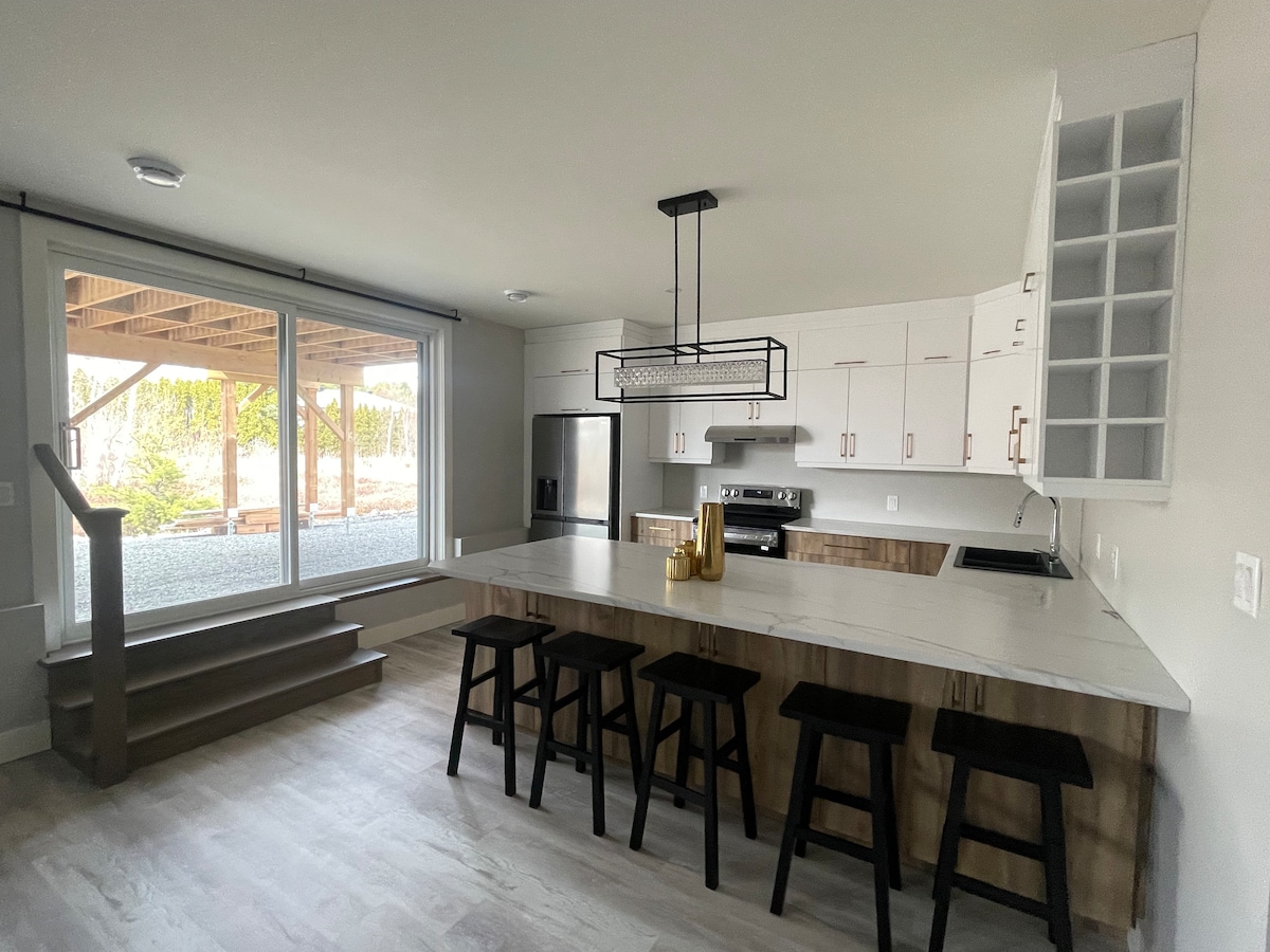 A modern kitchen features a large island with seating for several guests. Light wood cabinets and stainless steel appliances are present. A large window looks out to a covered outdoor area, providing a view of the surrounding landscape and natural light.