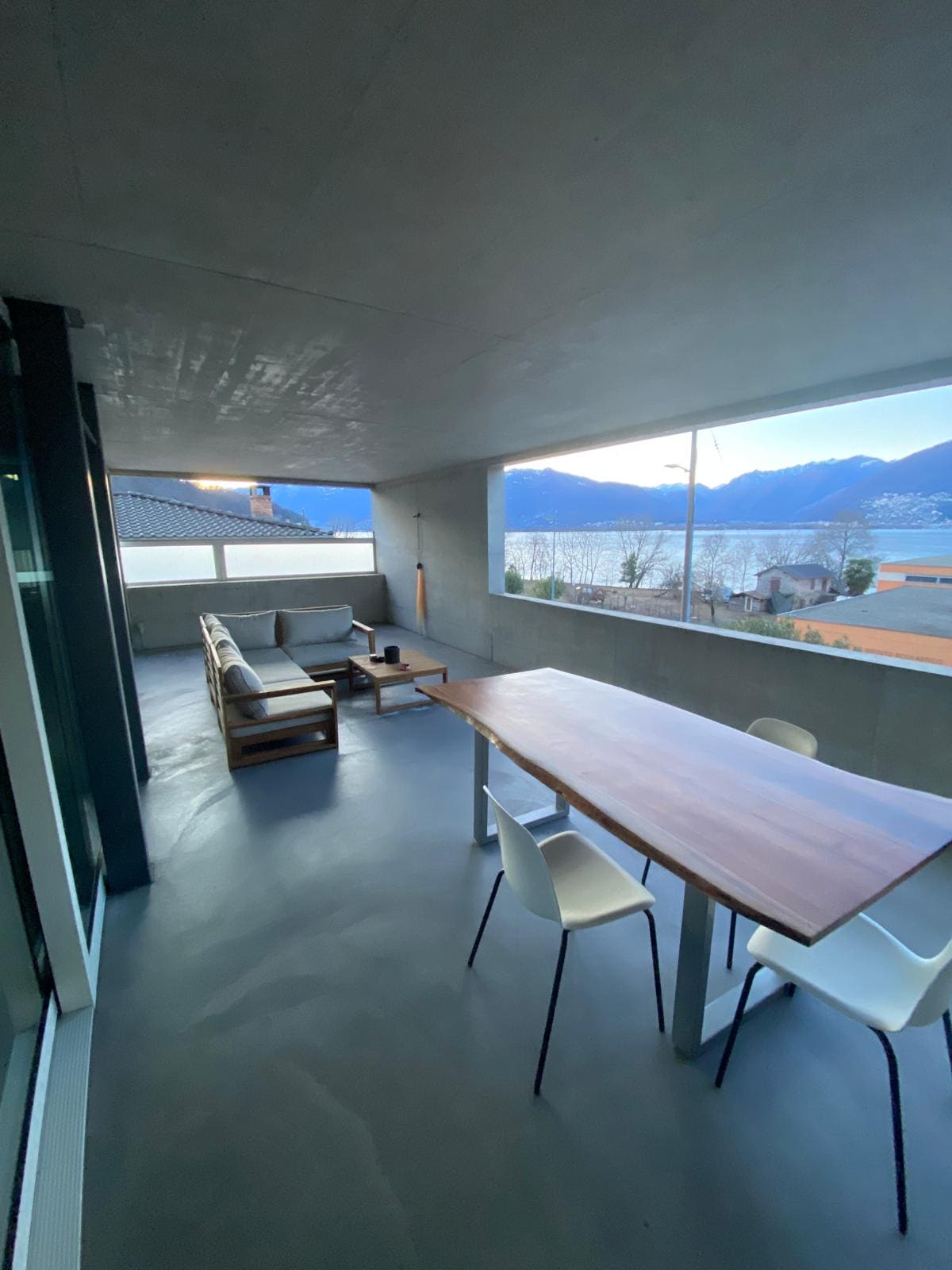 An expansive terrace is furnished with a comfortable outdoor sofa and a wooden dining table with chairs. Large windows frame views of the surrounding mountains and landscape, allowing natural light to fill the space. A serene and open atmosphere is created by the minimalist design.