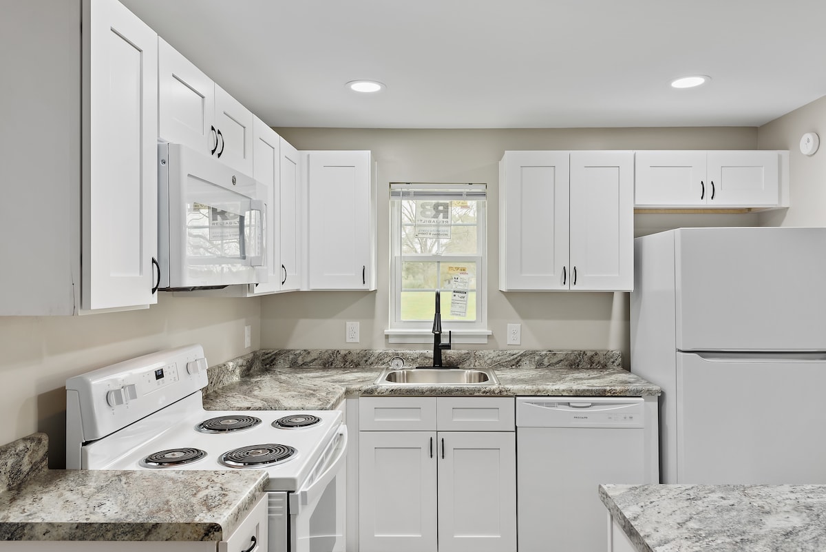 A well-equipped kitchen features clean white cabinetry, granite countertops, and essential appliances, including a stove with oven, microwave, and refrigerator. A double sink sits beneath a window, providing natural light, while ample cupboard space offers convenient storage.