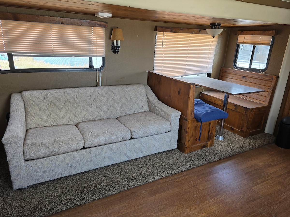 A light-colored couch with a textured pattern is positioned against a wall. A wooden dining table with two benches is adjacent, illuminated by natural light from nearby windows with blinds. The flooring is finished in warm wood tones, contributing to a cozy environment.