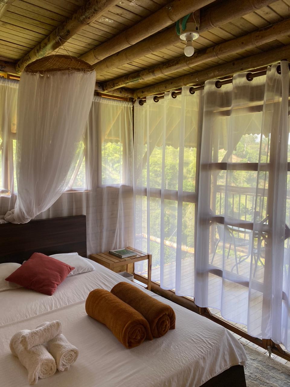 A bright and airy bedroom features a spacious bed with soft linens, accented by decorative pillows. Large sheer curtains allow natural light to fill the room while providing views of the greenery outside. A balcony with seating is visible through the glass doors.