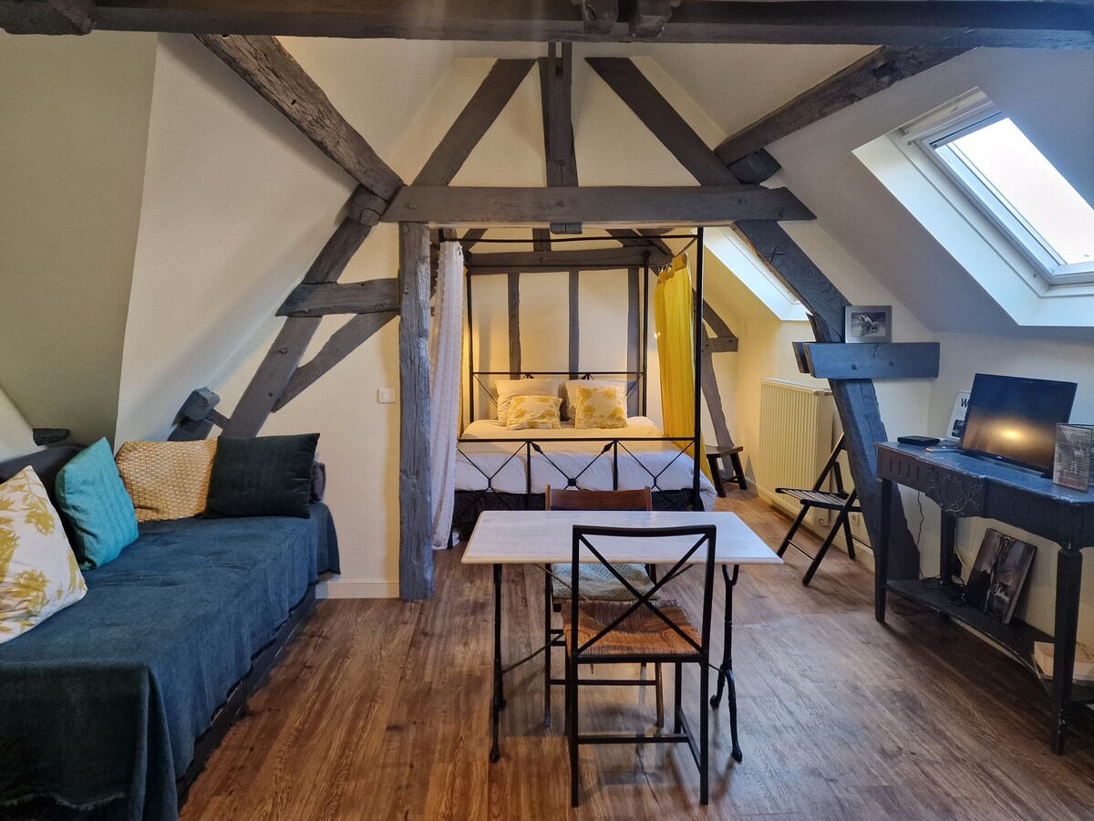 An attic room features exposed wooden beams and sloped ceilings. A spacious bed is framed by soft yellow curtains, complemented by a cozy seating area with a dark throw. A wooden table and chairs provide a functional dining space. Natural light fills the room through a skylight.