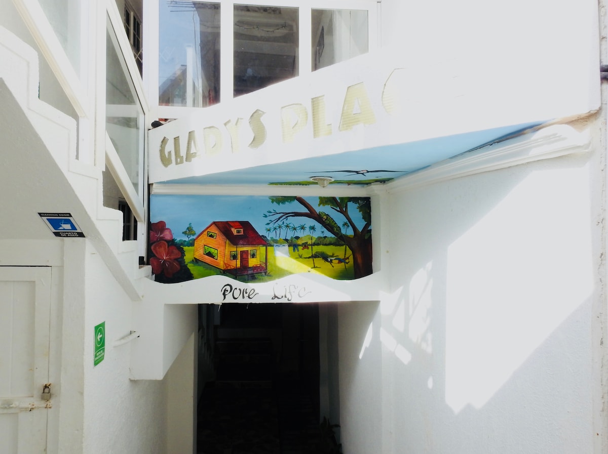 An entrance area is displayed, featuring a mural depicting a colorful house surrounded by trees. The words 'Gladys Place' are painted above, and a staircase leads into the building. Light filters into the space, creating a welcoming atmosphere for guests.