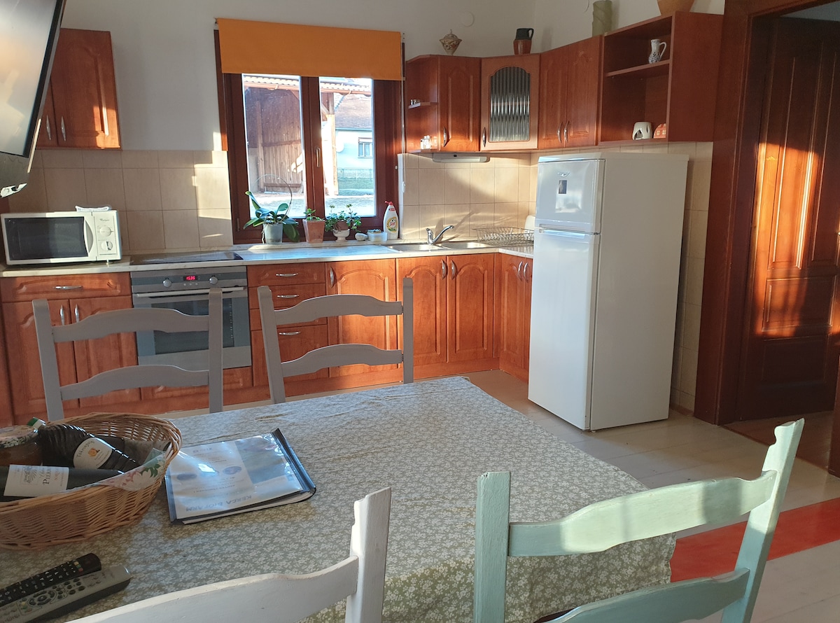 The kitchen is equipped with wooden cabinetry and ample countertop space. A refrigerator, oven, and microwave are visible. A dining table is surrounded by light-colored chairs, with a basket on the table. Light enters through a window covered by a bright orange curtain.