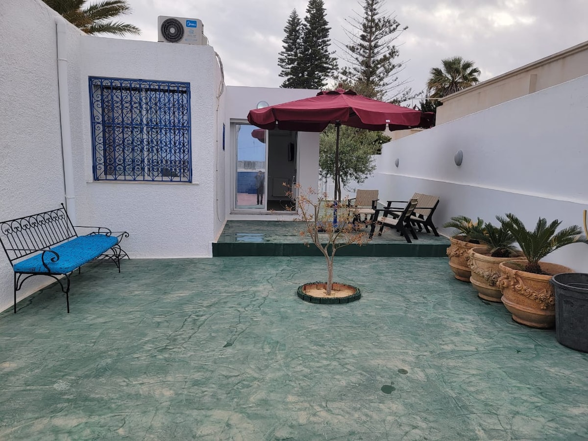 An outdoor area is presented with green stamped concrete flooring. A shaded area is provided by a maroon umbrella, with a small table and chairs beneath it. Potted plants are arranged around the space, while a blue bench offers additional seating.