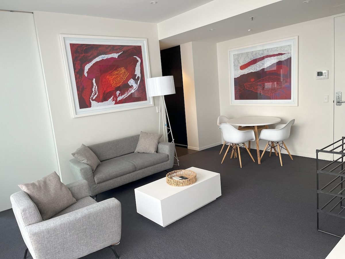 A modern living area features a light gray sofa and an armchair accompanied by a white coffee table. Two vibrant abstract artworks adorn the walls, while a small dining table with white chairs is positioned nearby, all set against a neutral color palette.
