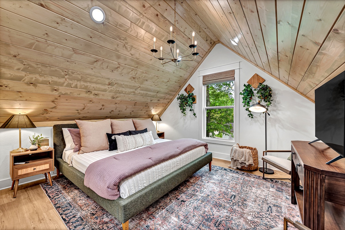 A spacious bedroom features a sloped wooden ceiling and large windows framed by greenery. A comfortable bed is adorned with decorative pillows and a lightweight blanket. A stylish chandelier hangs above, complementing the modern decor, while a rug adds texture beneath the furniture.