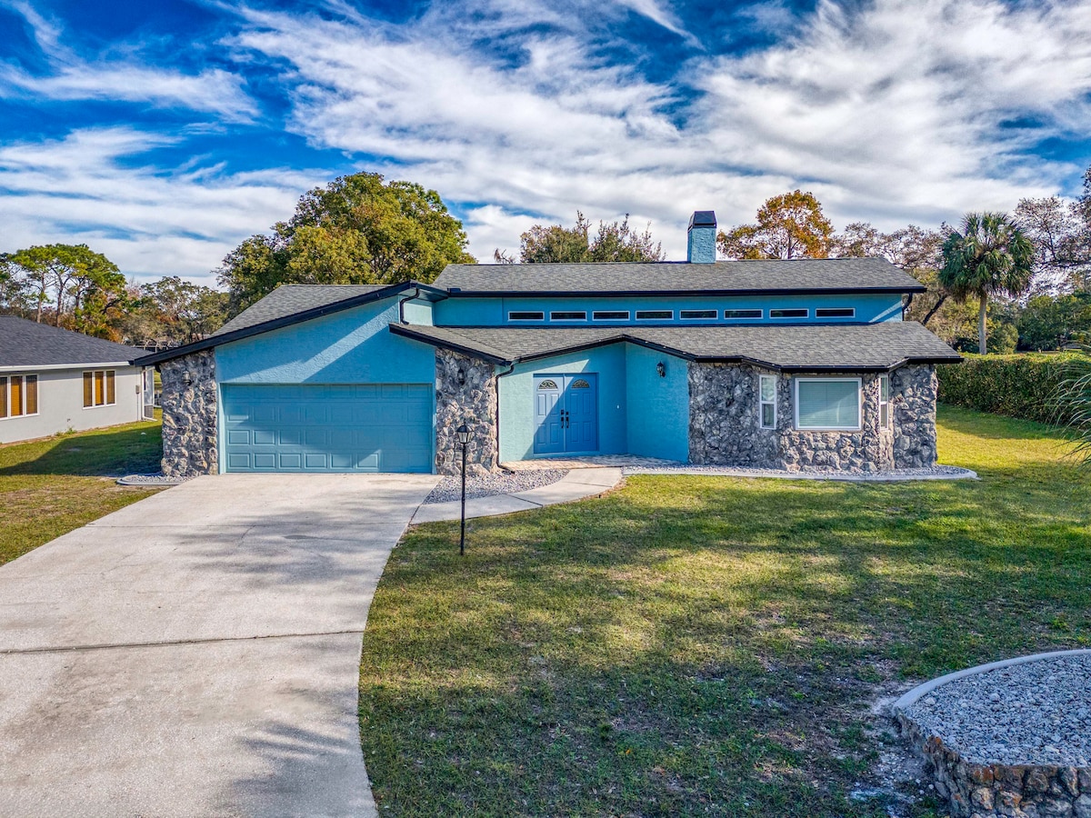 A modern home exterior is presented, showcasing a striking blue facade complemented by stone accents. The driveway curves towards a spacious entrance, framed by well-maintained lawns and trees. A clear blue sky with scattered clouds serves as a backdrop.