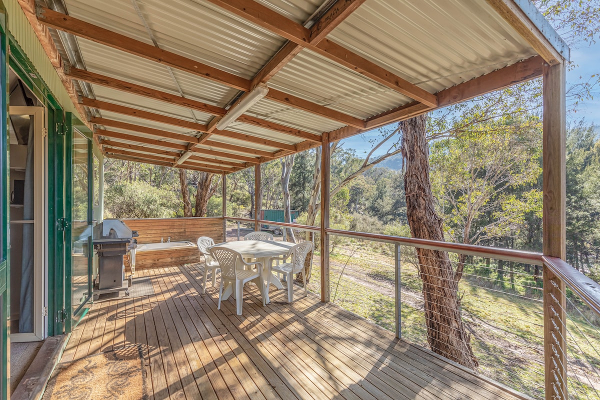 A covered deck features a wooden floor, surrounded by trees and natural greenery. A round table with plastic chairs is positioned for dining, and a barbecue station stands ready for outdoor cooking. Natural light filters through the space, enhancing the serene setting.